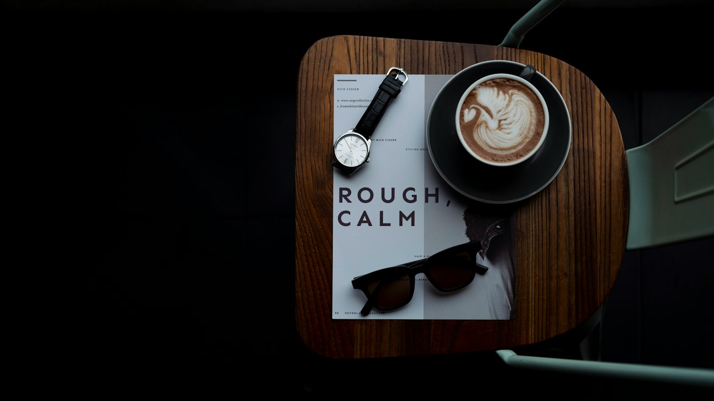 Zegarek, okulary przeciwsłoneczne, magazyn i latte art na drewnianym stole.